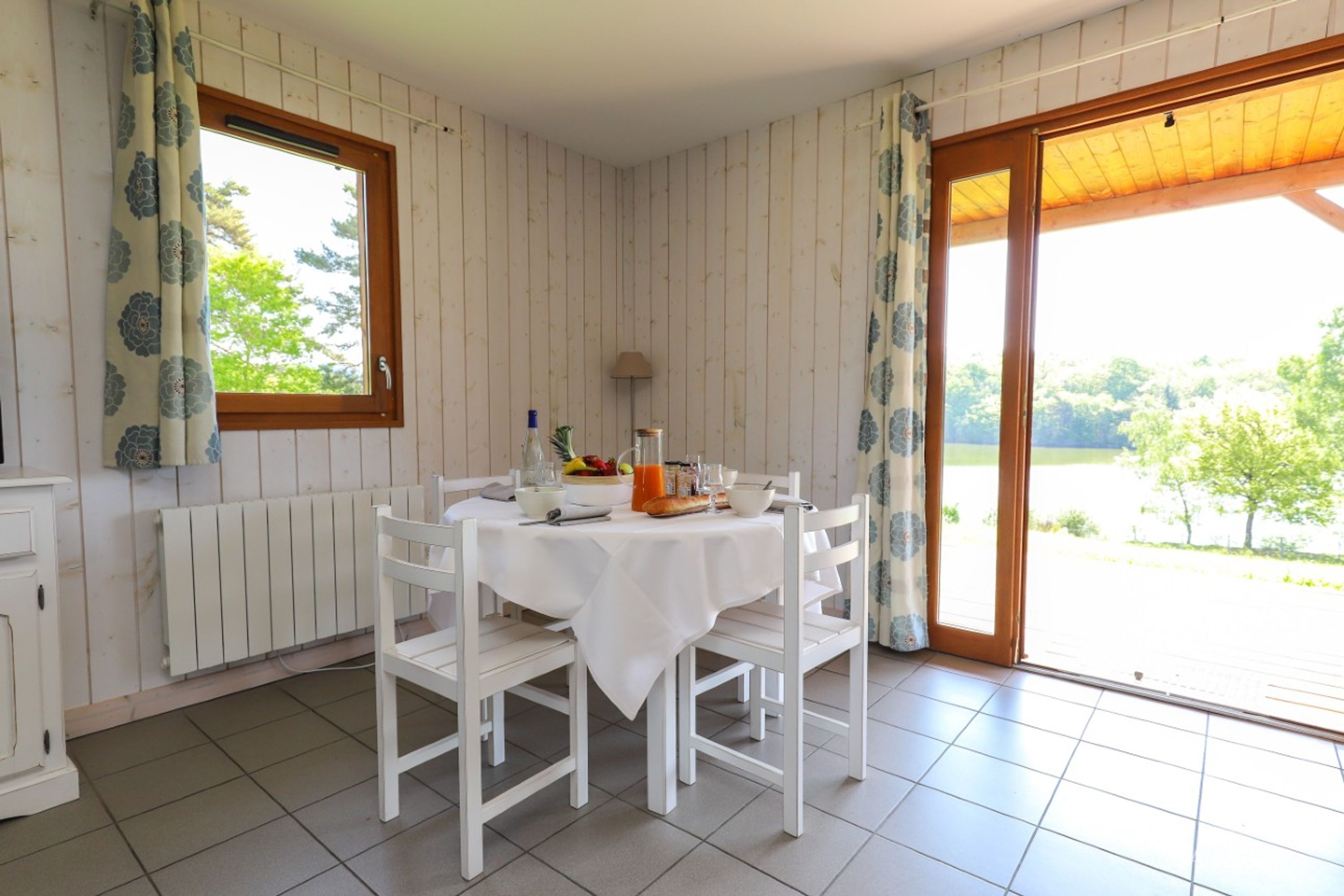 Un comedor con una mesa y sillas blancas, una ventana con cortinas, un radiador y vistas al exterior con vegetación.