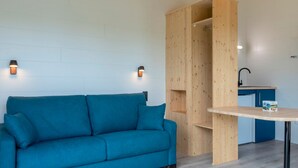 A modern room with a teal sofa, wooden shelving, a small table, and a kitchen area. - Le Domaine des Mon di res 1p4v (Meyrignac-l'Eglise)