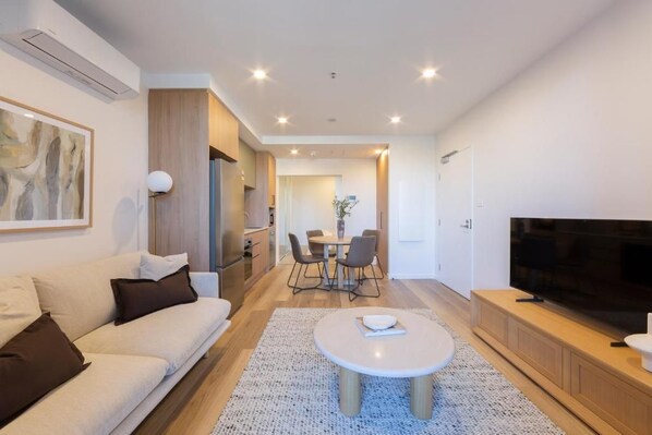 A modern living room with a sofa, coffee table, TV, and dining area. - Canopy Collective Serviced Apartments (Phillip)