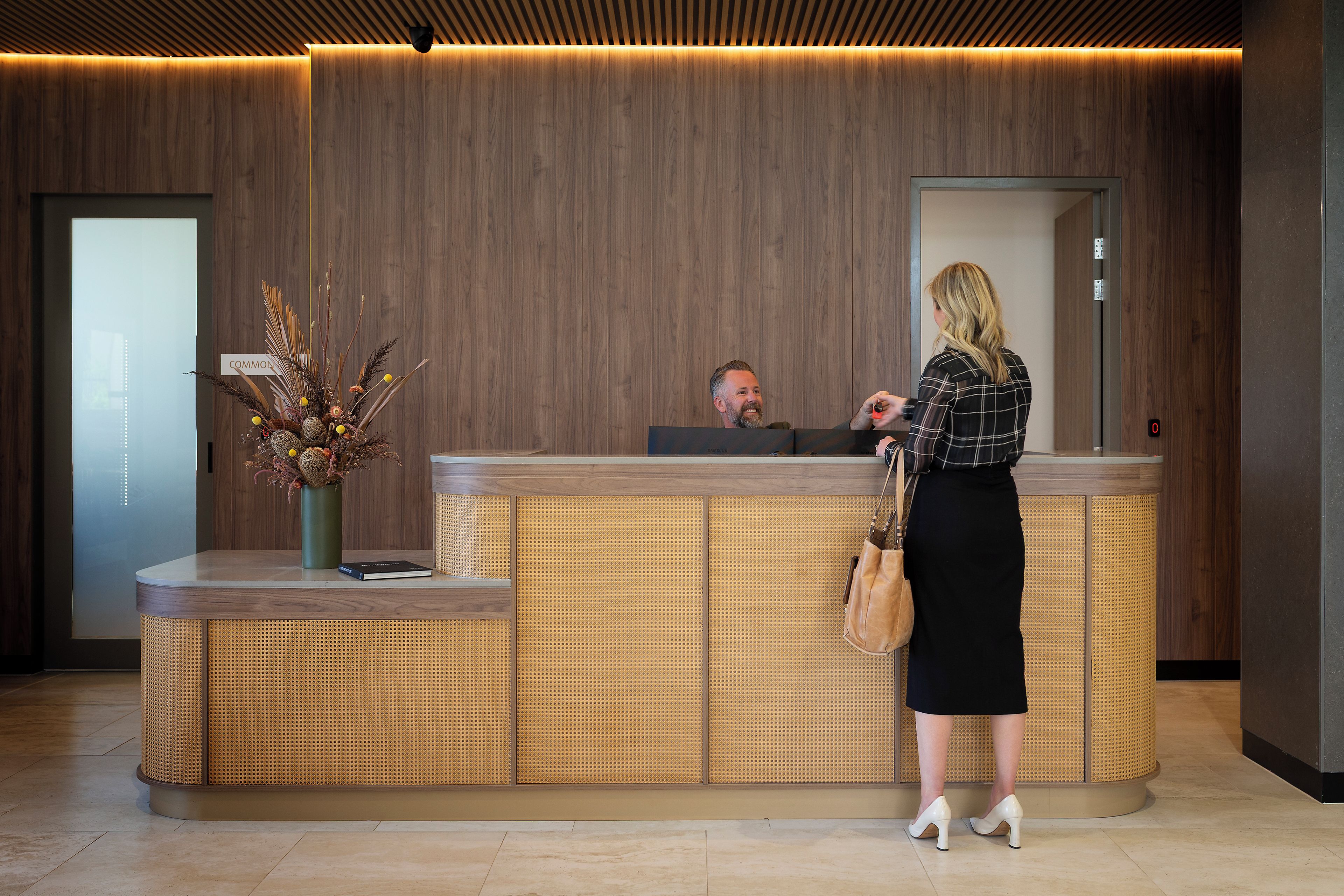 A reception desk with a wooden panel backdrop, a vase with dried flowers, and a person standing at the counter.