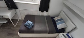Quarto pequeno e bem organizado, com uma cama, uma escrivaninha e uma cadeira.