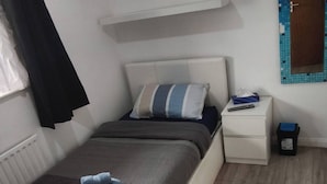 A bedroom with a bed, bedside table, and a small trash bin. - Room-small Cozy Room in Chelmsford Sprin (Chelmsford)