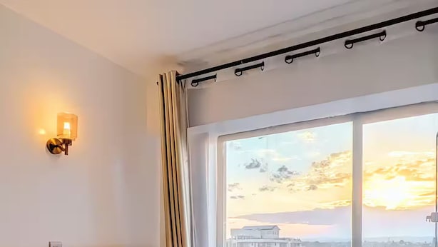 Une chambre d’hôtel avec un grand lit, une tête de lit, une lampe fixée au mur et une vue sur le paysage urbain, visible à travers une grande fenêtre.