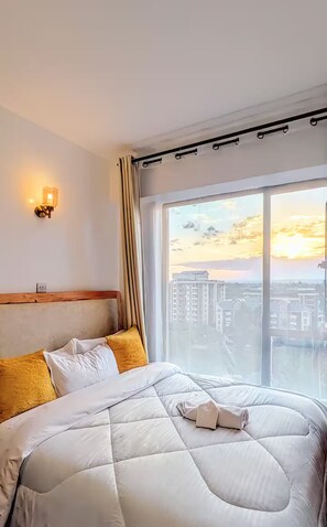 A hotel room with a large bed, a headboard, a wall-mounted light, and a view of a cityscape through a large window. - Pearl Executive Studio • Access UN Gigiri & Karura • Mountain Views • Fast Wi-Fi (Kiambu Province)