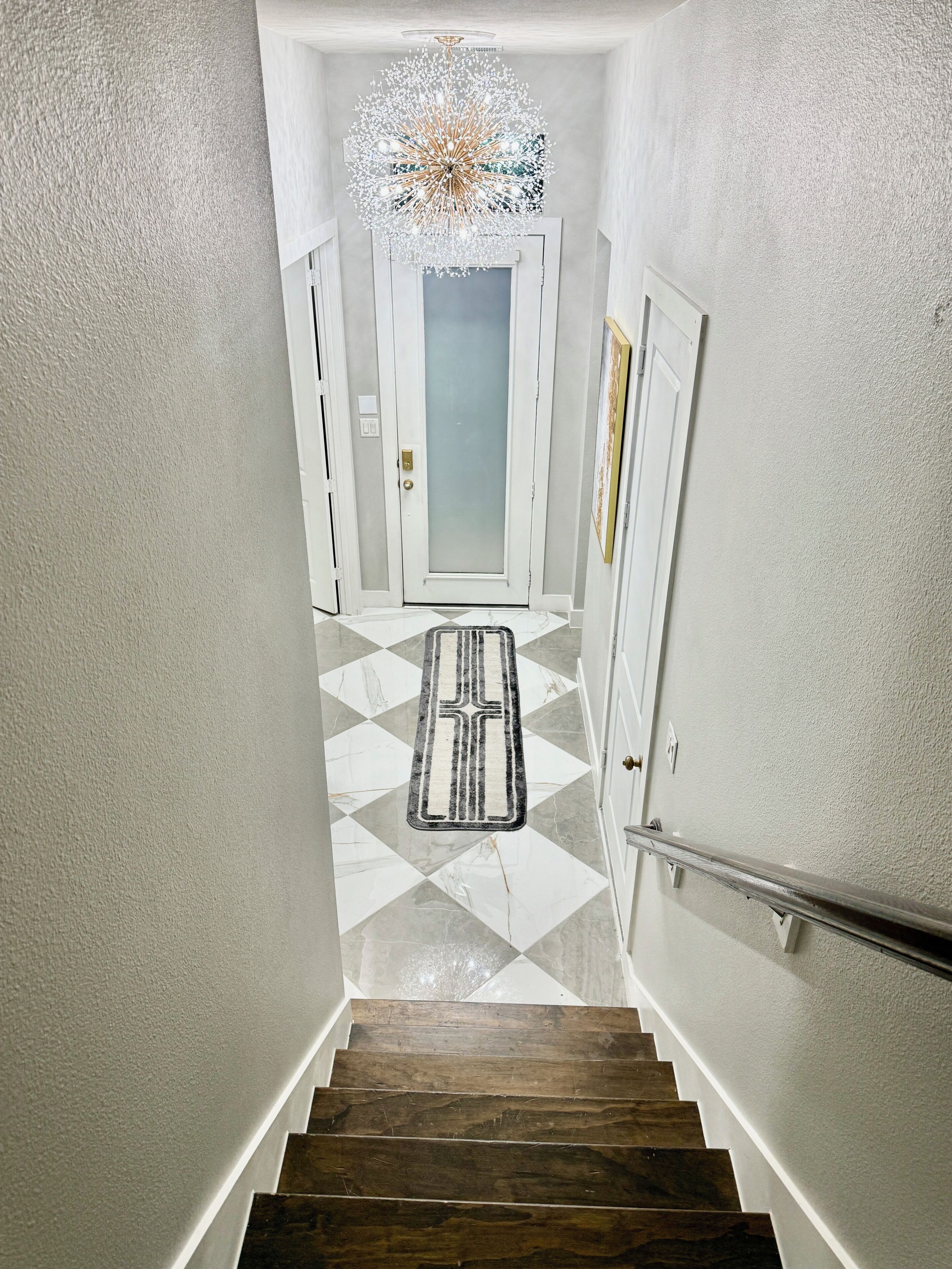 A staircase leading to a door with a geometric patterned floor, a modern chandelier, and a white door with a frosted glass panel.