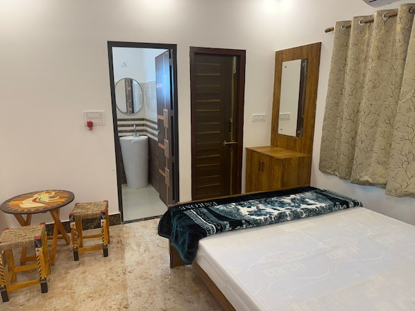 A bedroom with a bed, a wooden wardrobe, a small table with stools, a mirror, and a bathroom visible through an open door.
