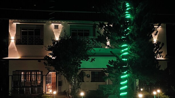 A two-story building with a green-lit spiral structure in front, surrounded by evenly spaced street lamps. - Jonaki Shantiniketan (Bolpur)