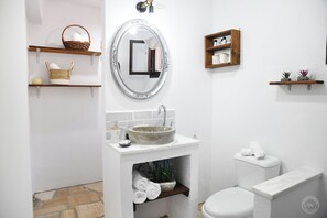 Casa de banho com lavatório branco, espelho redondo, sanita e prateleiras com toalhas e artigos de higiene pessoal.