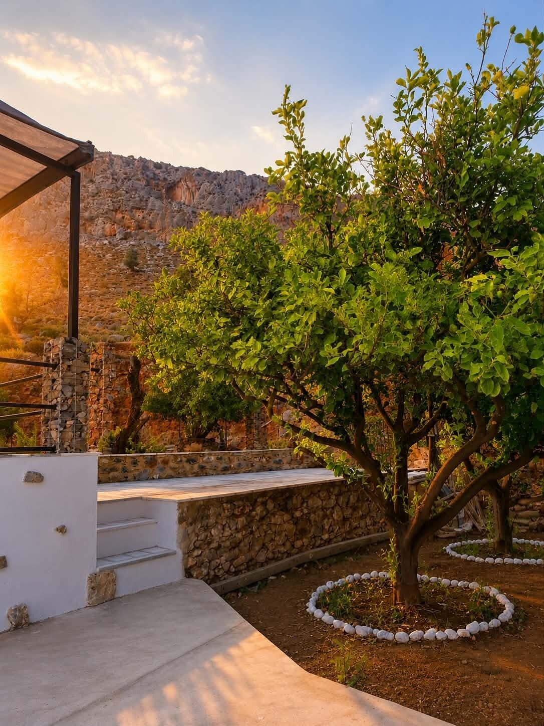 Un muro de piedra con una estructura de pérgola, un árbol con un borde circular de piedra y montañas al fondo.
