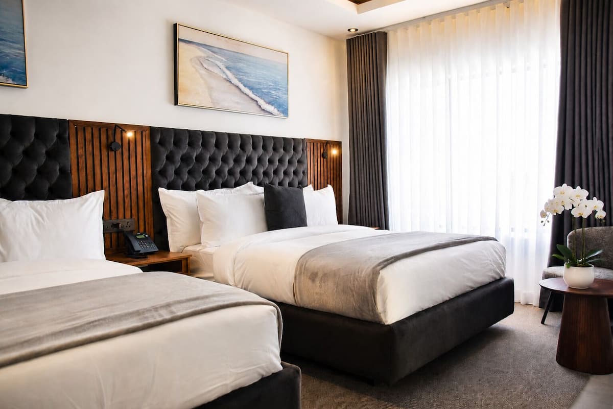 A hotel room with two beds, a large headboard, a painting, a telephone, and a plant.