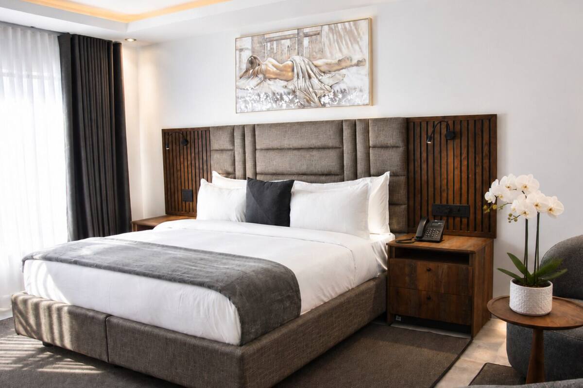 A modern hotel room with a large bed, a wooden headboard, a bedside table with a phone, and a small round table with a plant.