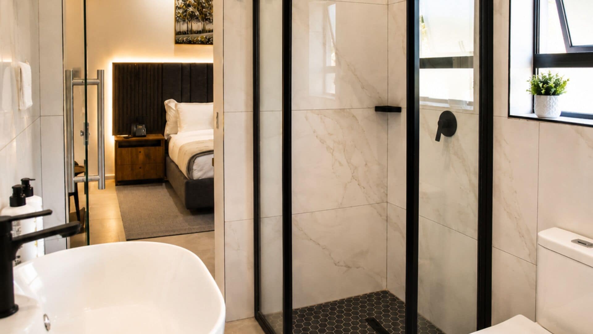Un baño moderno con cabina de ducha de vidrio, una bañera blanca y un dormitorio visible con cama y mesita de noche.