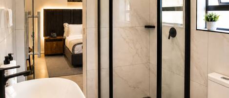Un baño moderno con cabina de ducha de vidrio, una bañera blanca y un dormitorio visible con cama y mesita de noche.