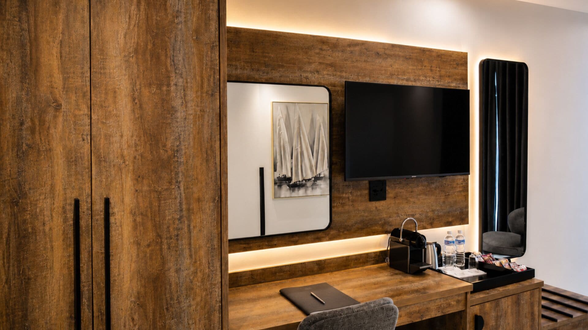 A modern hotel room with a wooden desk, a flat-screen TV, a framed artwork, and a small seating area.