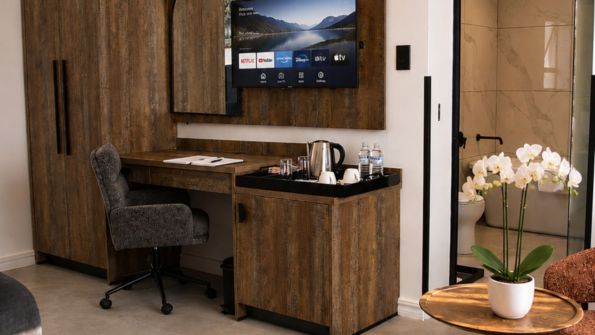 Un espacio de oficina moderno con un escritorio de madera, una silla, un televisor montado que muestra servicios de streaming y una orquídea en maceta.