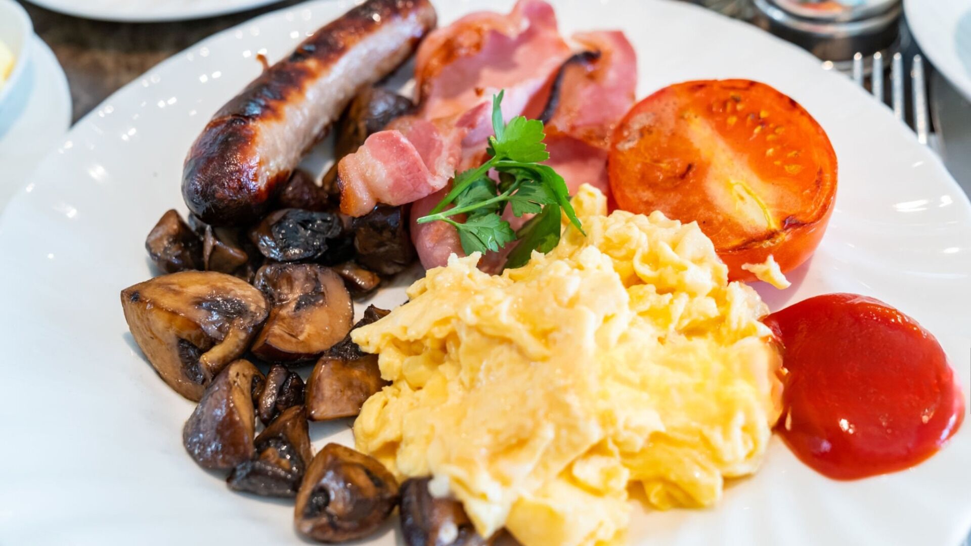 A plate of breakfast food including sausages, bacon, mushrooms, scrambled eggs, and a tomato.