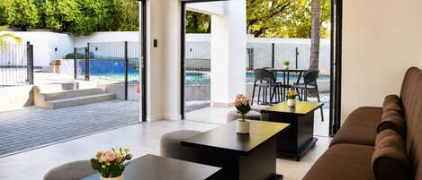 Una sala moderna con sofá, una mesa de centro y un comedor con sillas y una mesa. Hay un gran ventanal que da a un área exterior con piscina y sillas.