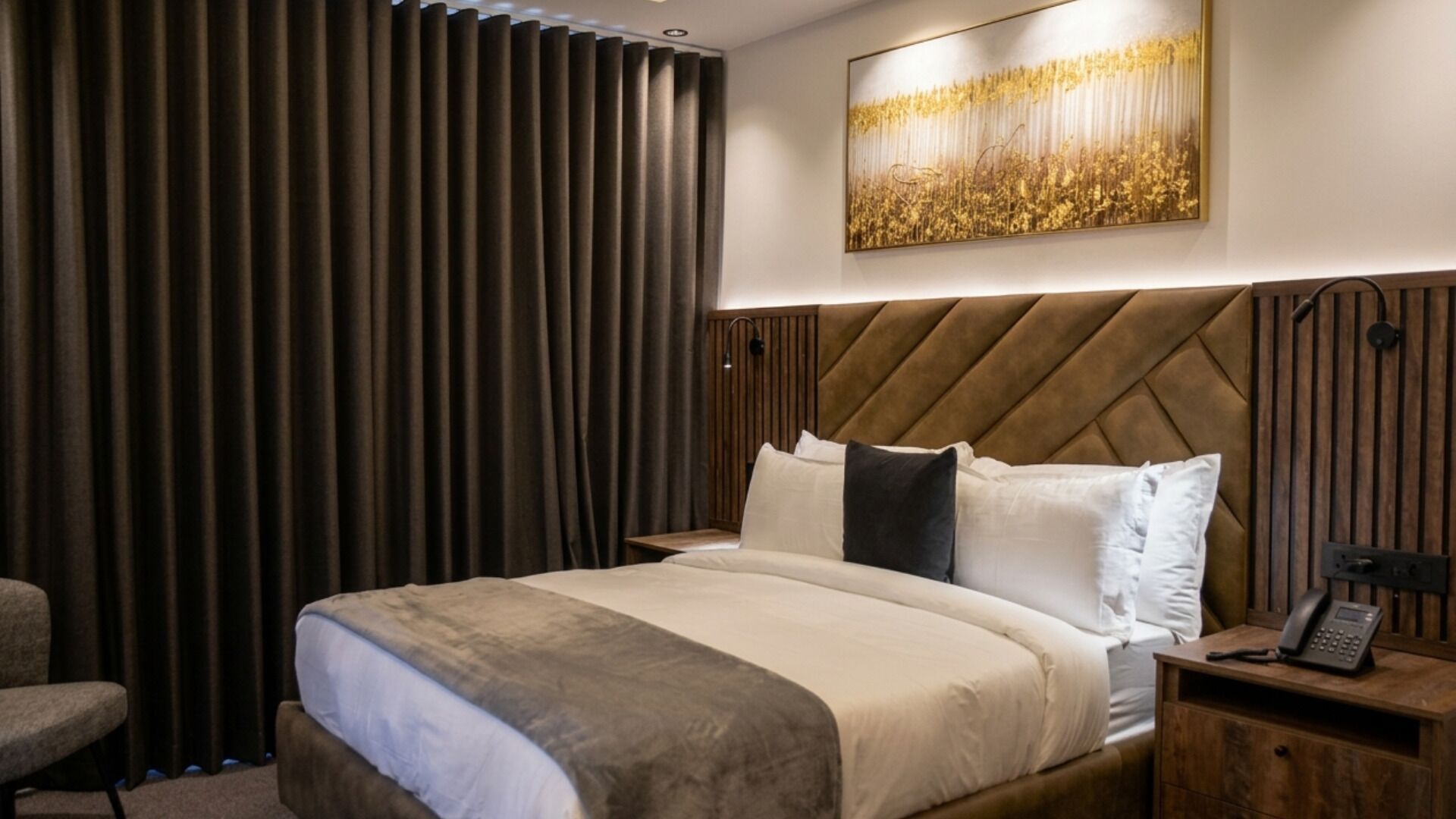 A hotel room with a bed, a nightstand, a chair, a large window with curtains, and a wall-mounted artwork.