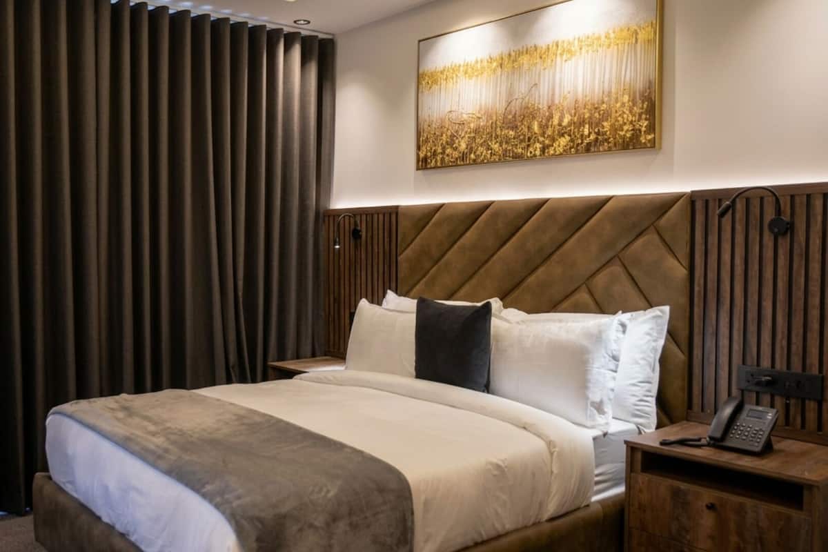 A hotel room with a bed, a nightstand, a chair, a large window with curtains, and a wall-mounted artwork.