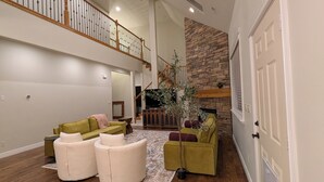 A spacious living room with a high ceiling, a staircase, a stone fireplace, and a large window. - River House - Entire 6-bedroom, 3.5 bathroom home near the SF River trail (Spanish Fork)