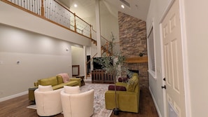 A spacious living room with a high ceiling, a staircase, a stone fireplace, and a large window.