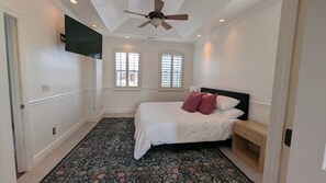 A bedroom with a bed, a ceiling fan, a TV, and two windows.