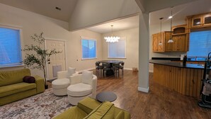 A living room with a sofa, ottomans, a dining table with chairs, a kitchen area with wooden cabinets, and a large window with blinds. - River House - Entire 6-bedroom, 3.5 bathroom home near the SF River trail (Spanish Fork)