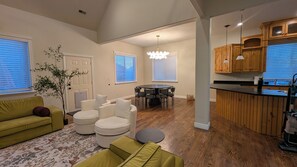 Un salon comprenant un canapé, des poufs, une table à manger avec des chaises, un coin cuisine avec des meubles en bois et une grande fenêtre avec des stores.
