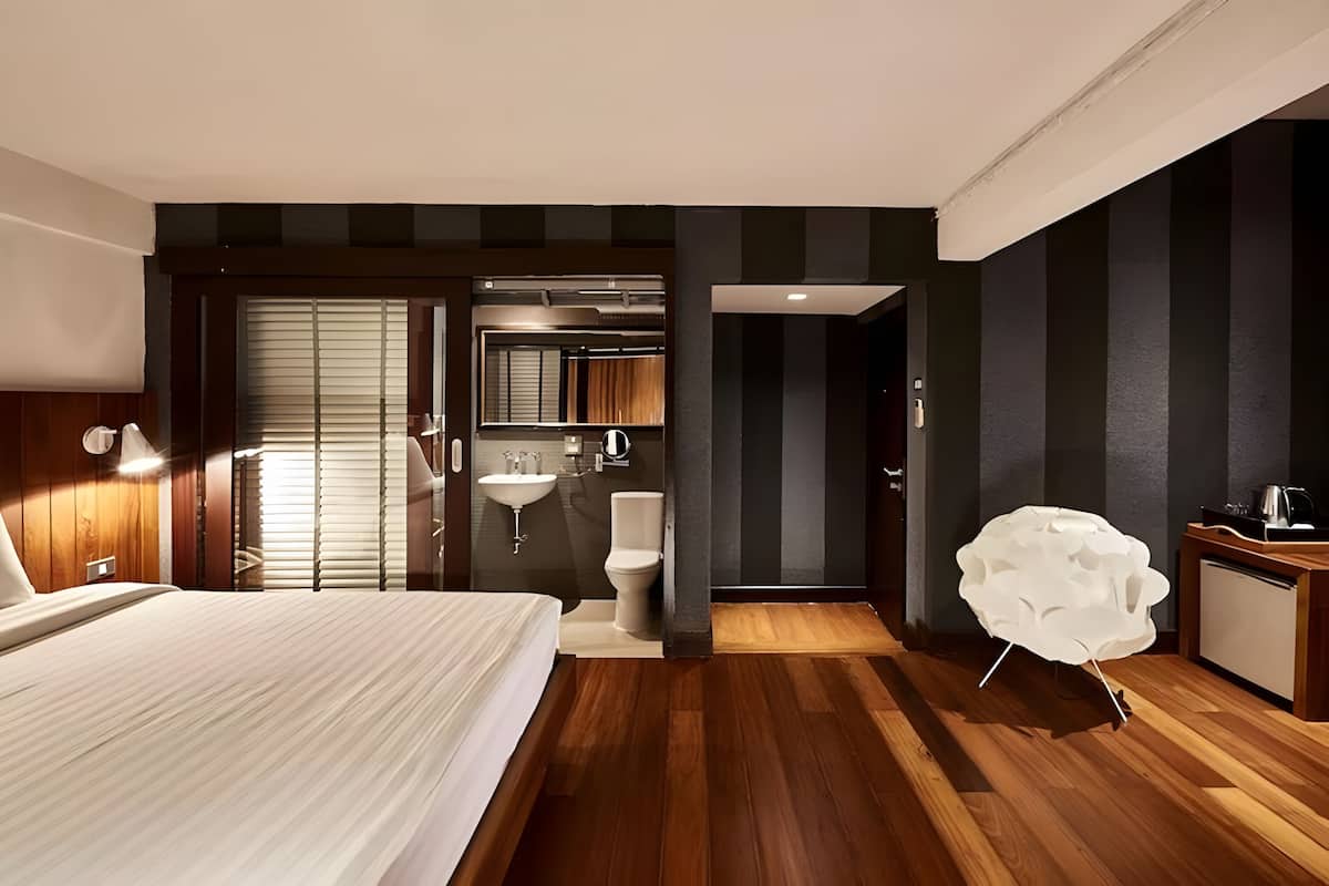 Une chambre d’hôtel moderne avec un grand lit, une salle de bain équipée d’un lavabo et de toilettes, et une chaise blanche au design original.