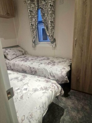 A small bedroom with two beds, a window with curtains, a wooden wardrobe, and a carpeted floor. - Marine Holiday Park - Holiday Accommodation 16604 (Rhyl)