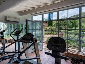 Ein Fitnessraum mit Laufbändern, einem Ergometer und einer Gewichtshebebank, mit großen Fenstern, die einen Blick auf einen Garten und Bäume bieten.