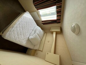 Ein kleines Zimmer mit einem Bett, einem Tisch, einem Fenster mit Jalousien und einer Wandleuchte.