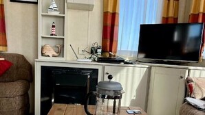 A cozy living area with a wooden table, two coffee mugs, a coffee pot, a television, and a bookshelf with decorative items. - Primrose Valley Holiday Park - Holiday Accommodation 1421 (Filey)