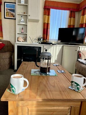 Ein gemütlicher Wohnbereich mit einem Holztisch, zwei Kaffeetassen, einer Kaffeemaschine, einem Fernseher und einem Bücherregal mit Dekoration.