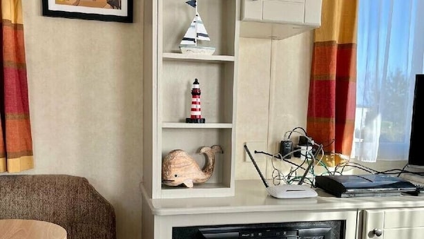 A living room with a fireplace, a framed picture of a coastal scene, a shelf with decorative items, and a window with curtains. - Primrose Valley Holiday Park - Holiday Accommodation 1421 (Filey)