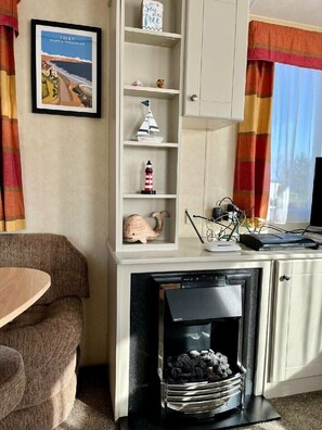 Ein Wohnzimmer mit Kamin, einem gerahmten Bild mit Küstenlandschaft, einem Regal mit Dekoration und einem Fenster mit Vorhängen.