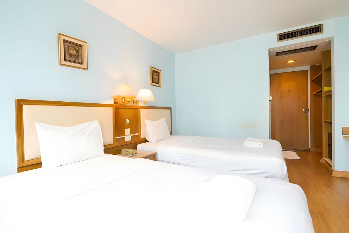 Habitación de hotel con dos camas, una mesita de noche con lámpara, un armario de madera y una puerta que da al baño.