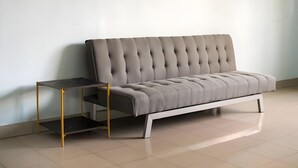 Ein modernes Sofa mit weißem Gestell und grauem gepolsterten Kopfteil, vor einer schlichten Wand in einem Raum mit Fliesenboden.