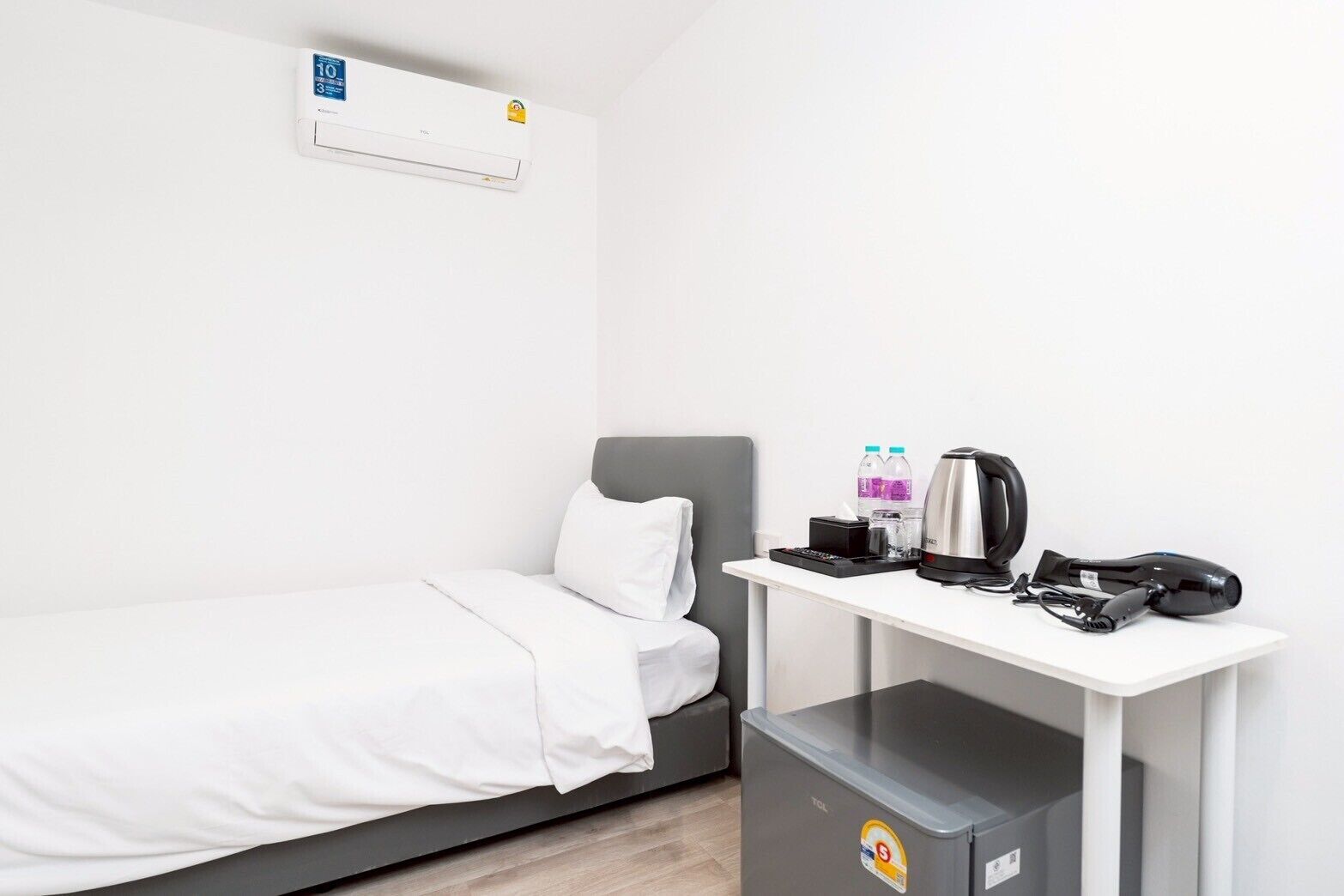 Habitación individual con cama blanca, cabecera gris, escritorio blanco con hervidor de agua, secador de pelo y una botella de agua.