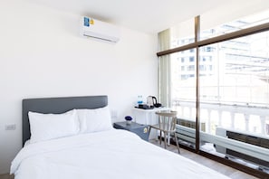 A hotel room with a bed, a small table with a kettle and water bottles, a chair, and a large window with a view of buildings.