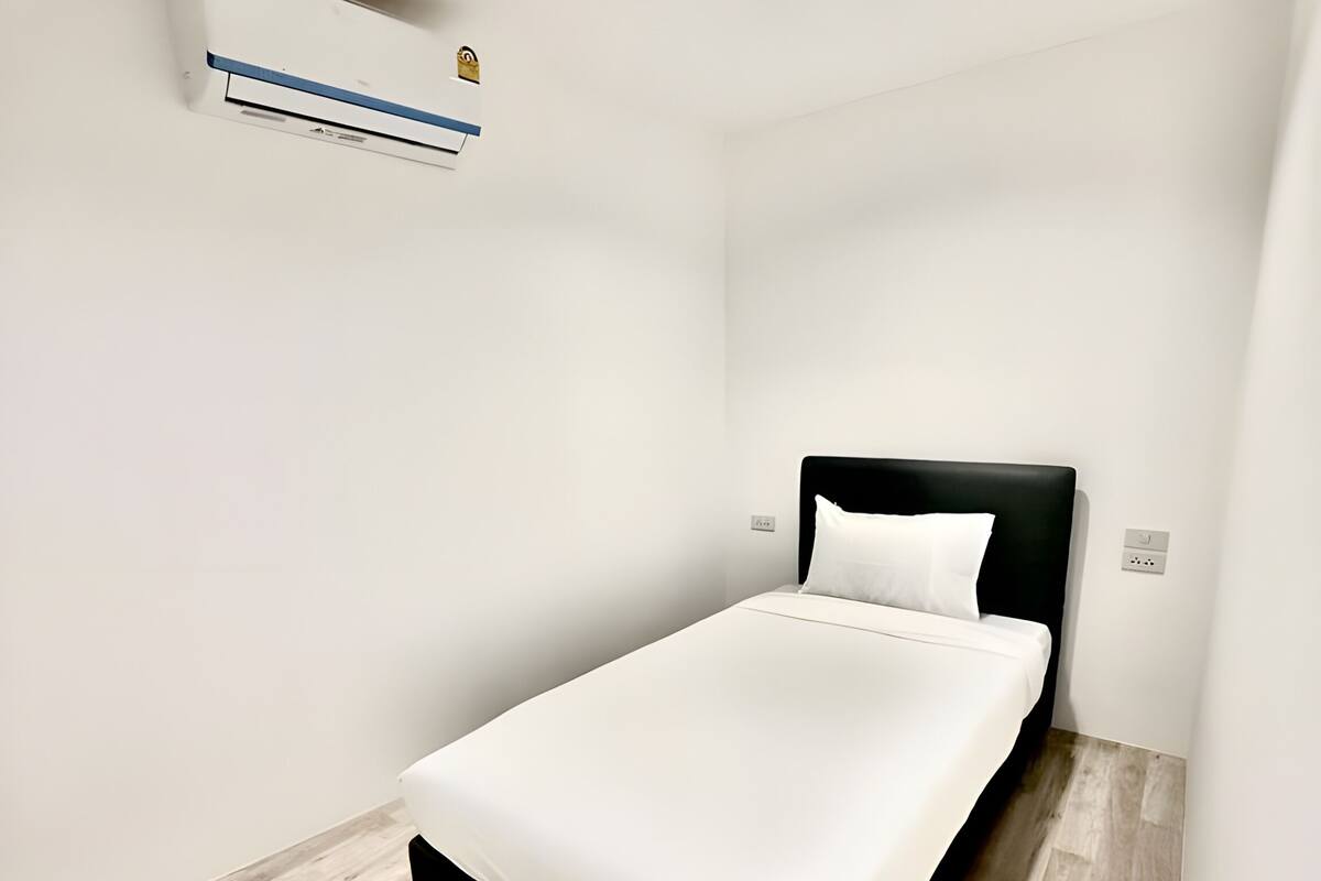 Una cama individual con cabecera negra y ropa de cama blanca en una habitación con suelo de madera clara y paredes blancas.