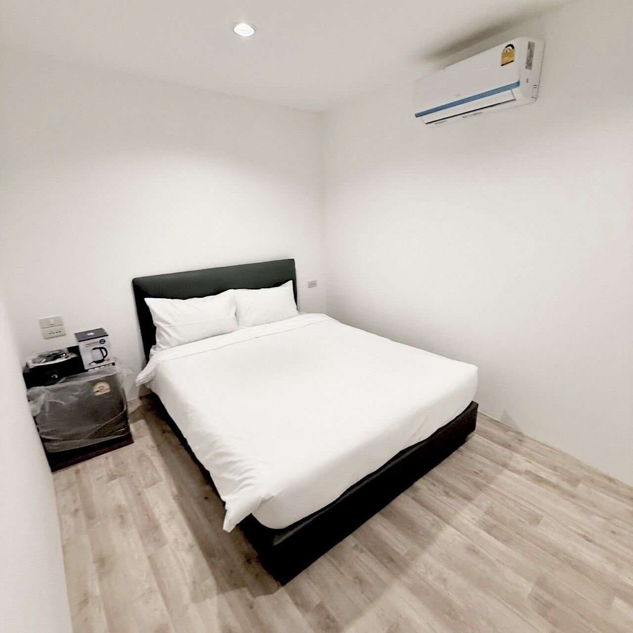 Ein kleines, sauberes Zimmer mit einem Einzelbett, einem Nachttisch, einem Mülleimer und einer Wand-Klimaanlage.