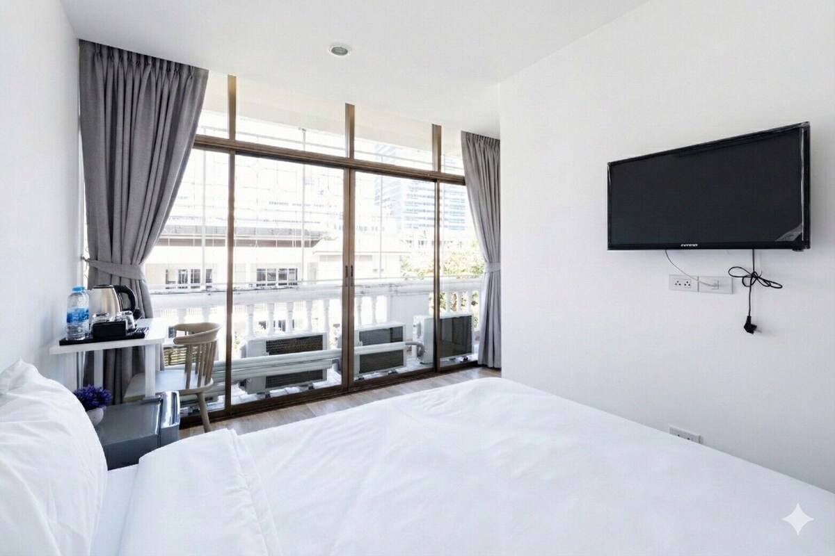 A hotel room with a bed, a desk with a kettle, a TV, and a balcony with a view of buildings.