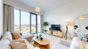 Ein modernes Wohnzimmer mit einer Couch, einem Fernseher und einer Lampe, großen Fenstern mit Vorhängen und Blick auf die Stadt.