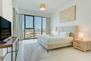 Ein Hotelzimmer mit einem großen Bett, einem Schreibtisch mit Fernseher, einem Nachttisch mit Lampe und Blick auf die Stadt durch das Fenster.
