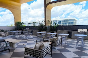 Een terras buiten met meerdere tafels en stoelen, een geruit vloertje en een uitzicht op een modern gebouw op de achtergrond.