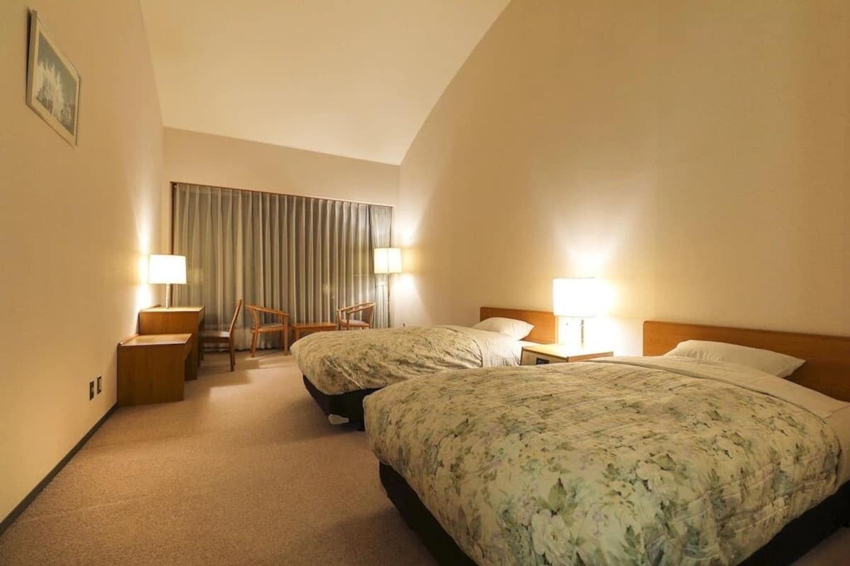 A hotel room with two beds, a desk, a chair, and a window with curtains.