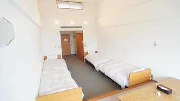 Ein Zimmer mit zwei Einzelbetten, einem Holztisch, einem kleinen Schreibtisch, einer Tür und einem Lüftungsgitter an der Wand.