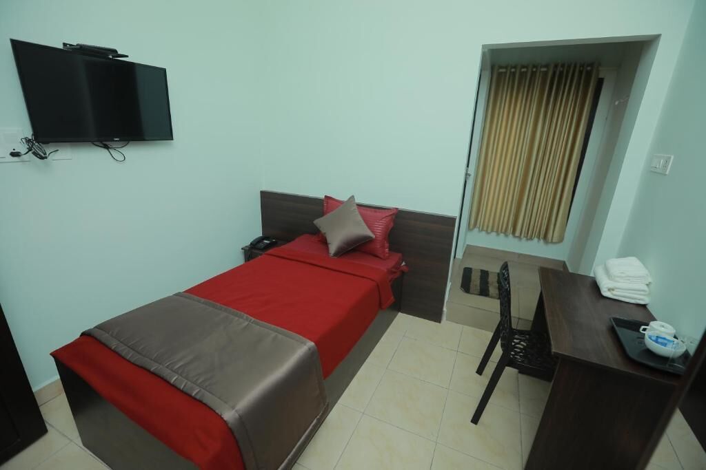 A single bed with a red and brown bedspread, a small desk with a chair, a television mounted on the wall, and a window with curtains.