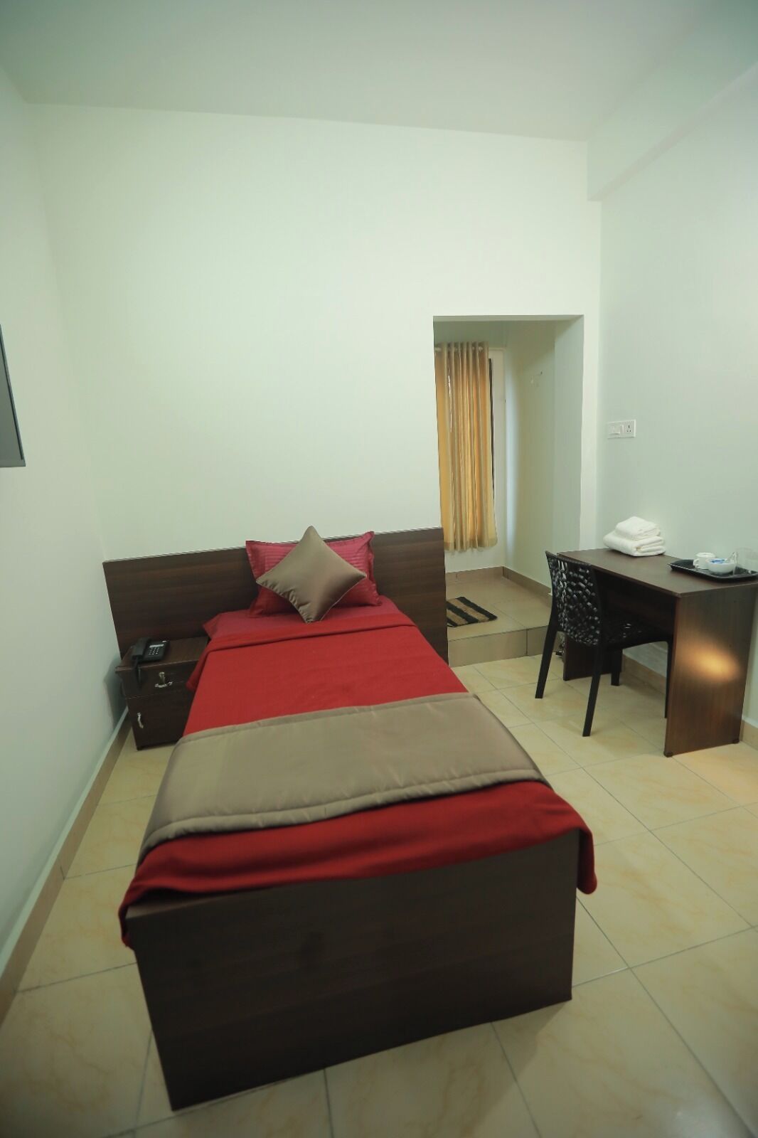 A compact hotel room with a bed, bedside table, desk, and a small window with curtains.